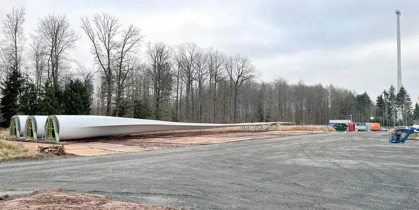 Zwei riesige, weiße Rotorblätter für Windkraftanlagen liegen auf einer Schotterfläche, daneben stehen Container und Fahrzeuge. Im Hintergrund ist ein Wald und ein Windturm zu sehen.