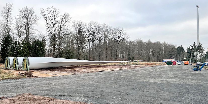 Zwei riesige, weiße Rotorblätter für Windkraftanlagen liegen auf einer Schotterfläche, daneben stehen Container und Fahrzeuge. Im Hintergrund ist ein Wald und ein Windturm zu sehen.