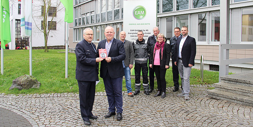 Eine Gruppe von Personen steht vor einem Gebäude mit EAM-Schild und hält ein Buch. Führende Positionen sind zu sehen.