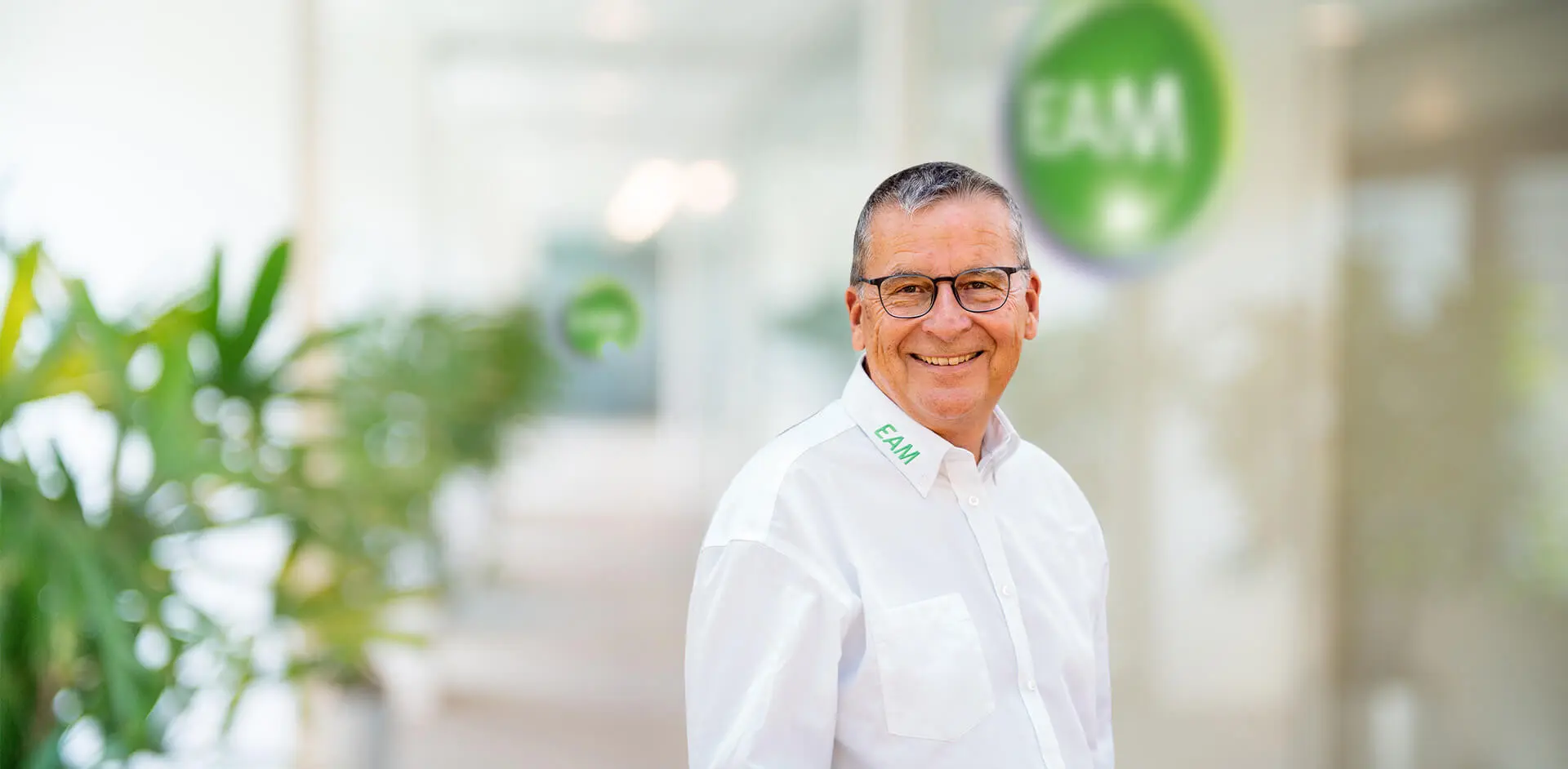 Ein lachender Mann mit Brille und Bart steht vor einem EAM-Logo in einem Büro.