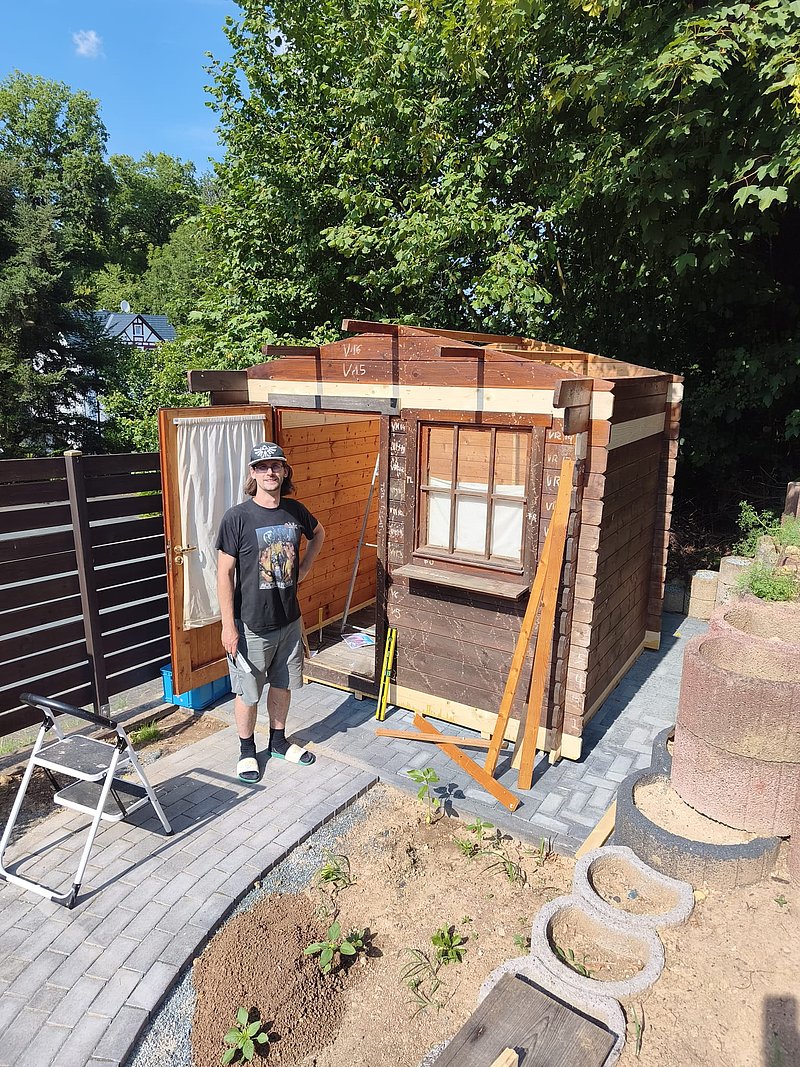 Person steht vor einem kleinen Holzhaus im Garten, umgeben von Pflanzen und Bäumen, bei sonnigem Wetter.