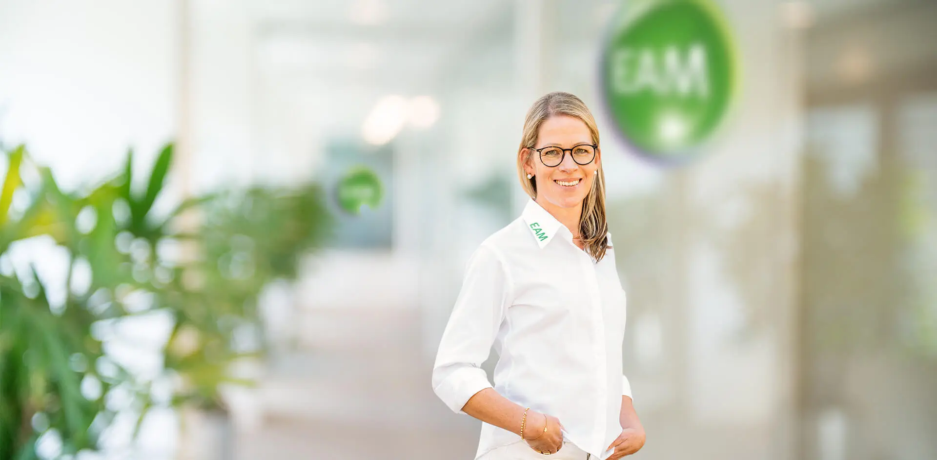 Eine lachende Frau steht vor einem EAM-Logo in einem Büro.