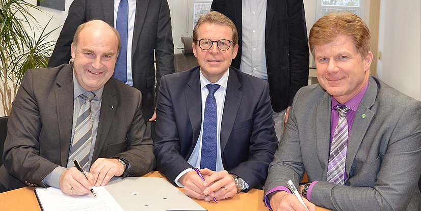 Drei Männer in Anzügen sitzen an einem Tisch und unterzeichnen Dokumente, lächelnd in einem Büroumfeld.