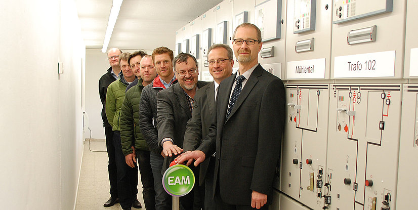 Gruppe von Männern in einem Technikraum, die gemeinsam einen Schalter betätigen, mit elektrischen Schaltanlagen im Hintergrund.