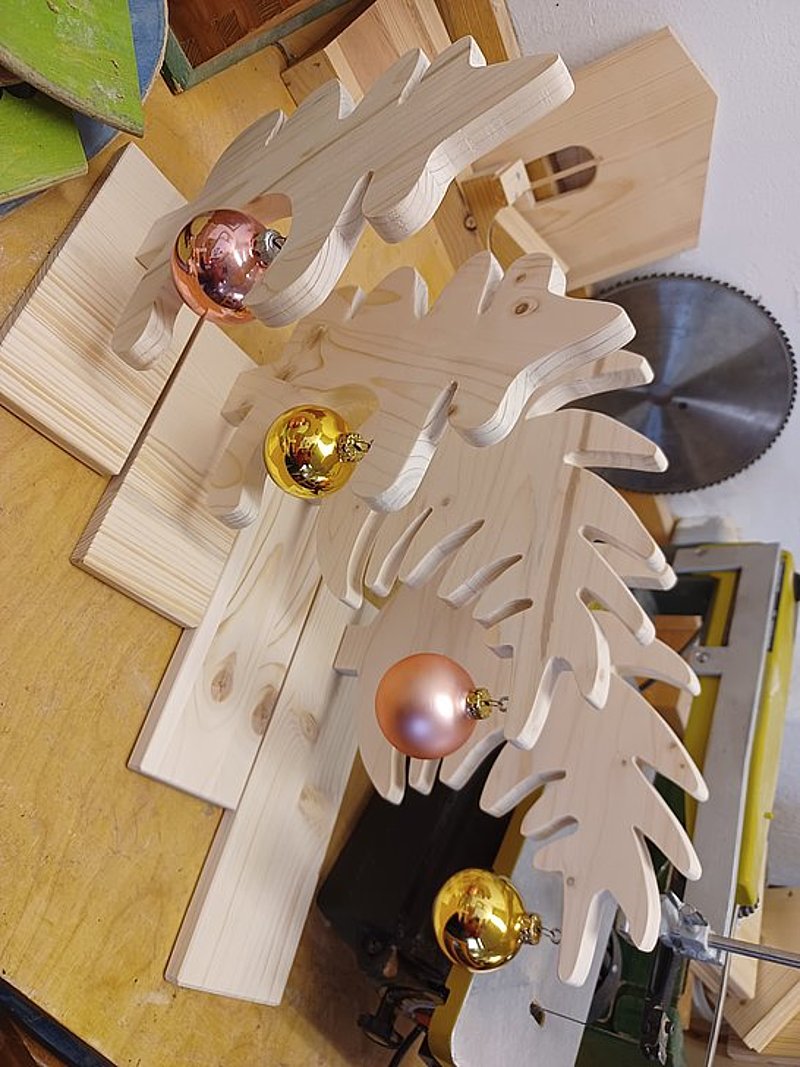 Dekorativer Holzbaum mit Weihnachtskugeln in Gold, Rosa und Orange auf einer Werkbank.