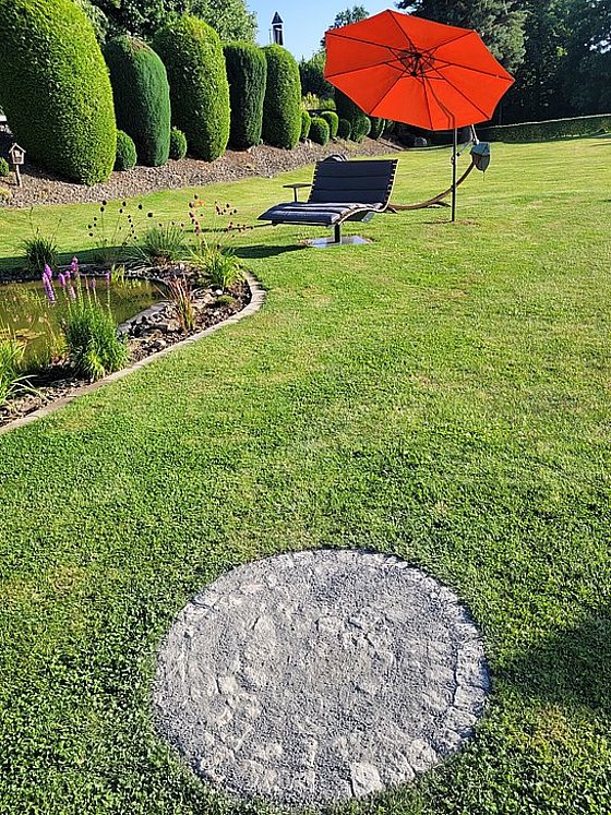 Sonnenschirm auf einer Liege im Garten, umgeben von Rasen und einem kleinen Teich mit Pflanzen.
