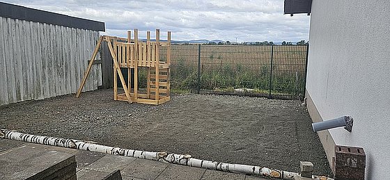 Ein leerer Gartenbereich mit einem Holzspielgerüst, umgeben von einer Zaunanlage und einem Blick auf die Landschaft.