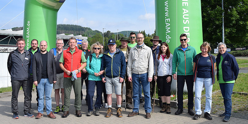 Gruppenfoto von 16 Personen vor einem grünen Bogen, umgeben von Bäumen und einem Gebäude im Hintergrund.