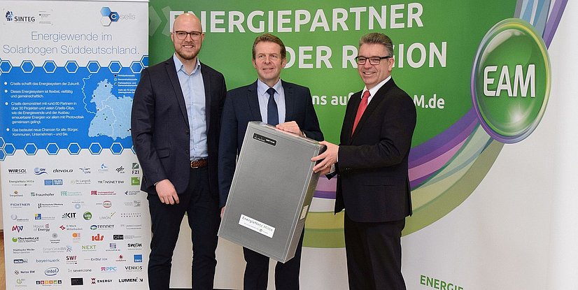 Drei Männer stehen vor einem Banner, der die Energiewende in Süddeutschland thematisiert, einer hält ein Gerät in der Hand.
