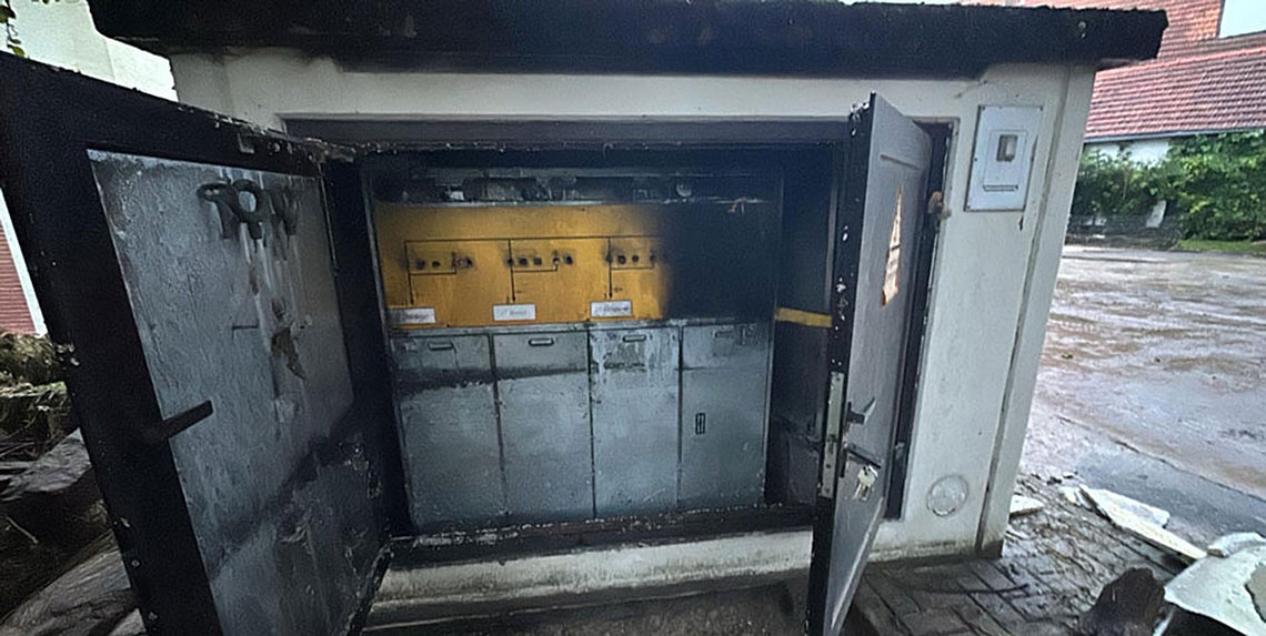 Eine graue, beschädigte Transformatorenstation mit weit geöffneten Türen zeigt im Inneren ein teilweise verkohltes gelbes Bedienfeld über grauen Schränken.
