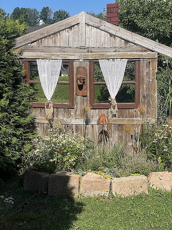 Holzhütte mit Fenster, Vorhängen, umgeben von Blumen und Pflanzen im Garten, Steinmauer im Vordergrund.