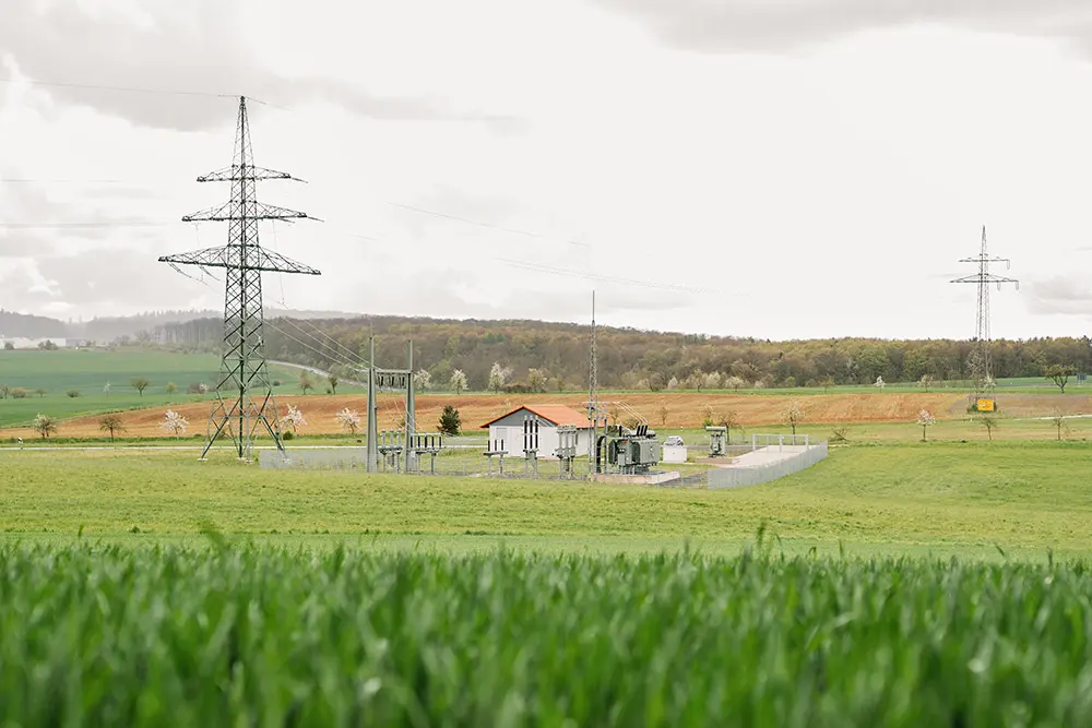 Ein Umspannwerk umgeben von Wiesen und hügeliger Landschaft, mit hohen Strommasten und einem bewölkten Himmel.