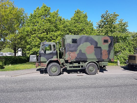 Ein militärischer Lastwagen steht auf einer Straße, umgeben von grünen Bäumen und klarem blauem Himmel.