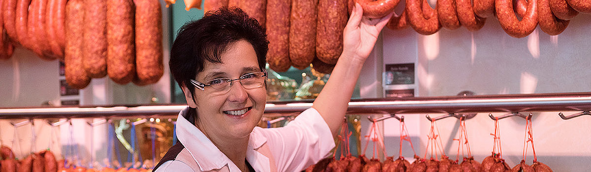 Frau in einem Feinkostladen lächelt und zeigt auf eine Wurstschlange, umgeben von verschiedenen anderen Wurstwaren.