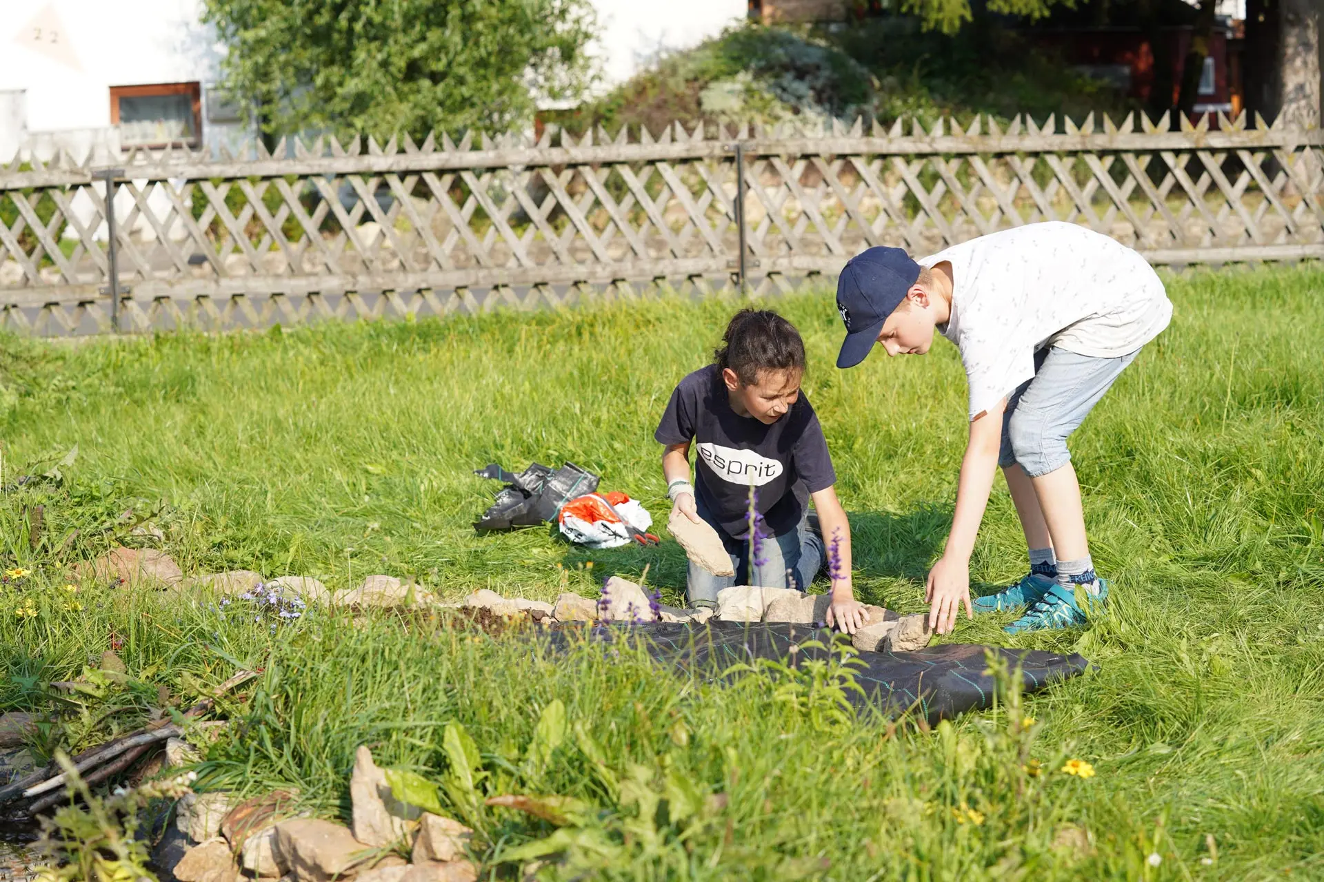 Zwei Kinder im Garten legen Steine auf eine schwarze Folie, um eine Fläche zu gestalten, umgeben von grünem Gras.