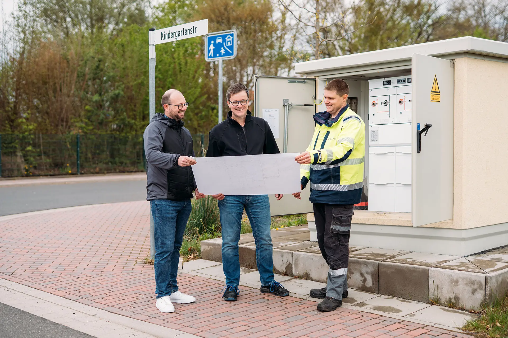 Drei Männer stehen neben einer technischen Anlage im Freien und betrachten gemeinsam einen großen Plan; im Hintergrund ist ein Straßenschild mit der Aufschrift „Goethestr.“ zu sehen.
