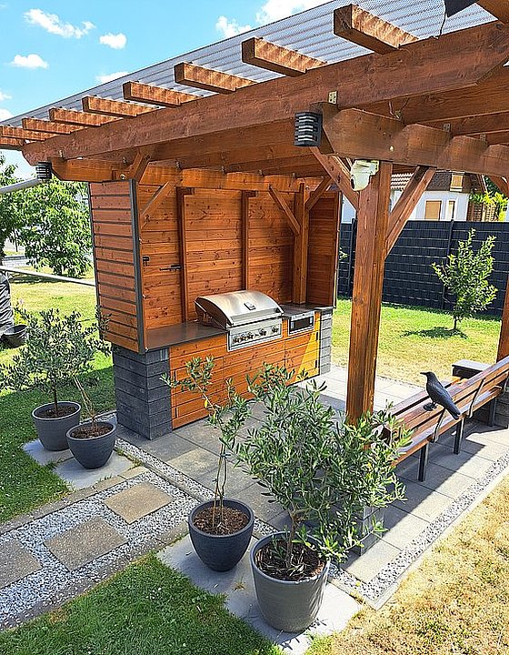 Holzkonstruktion mit Grill, umgeben von Pflanzen und einer Sitzbank in einer grünen Gartenlandschaft.