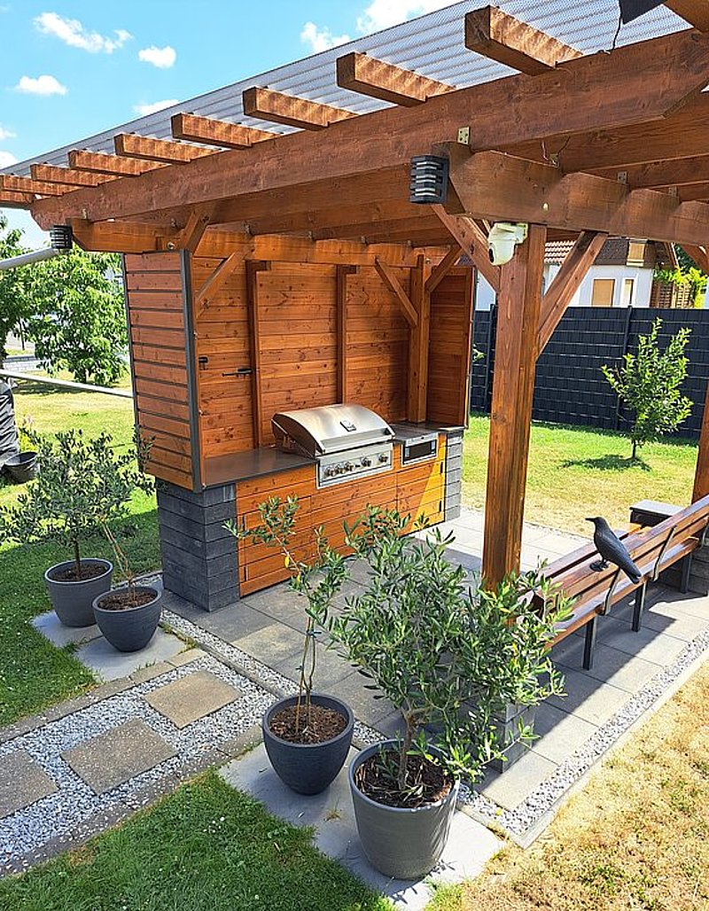 Holzkonstruktion mit Grill, umgeben von Pflanzen und einer Sitzbank in einer grünen Gartenlandschaft.