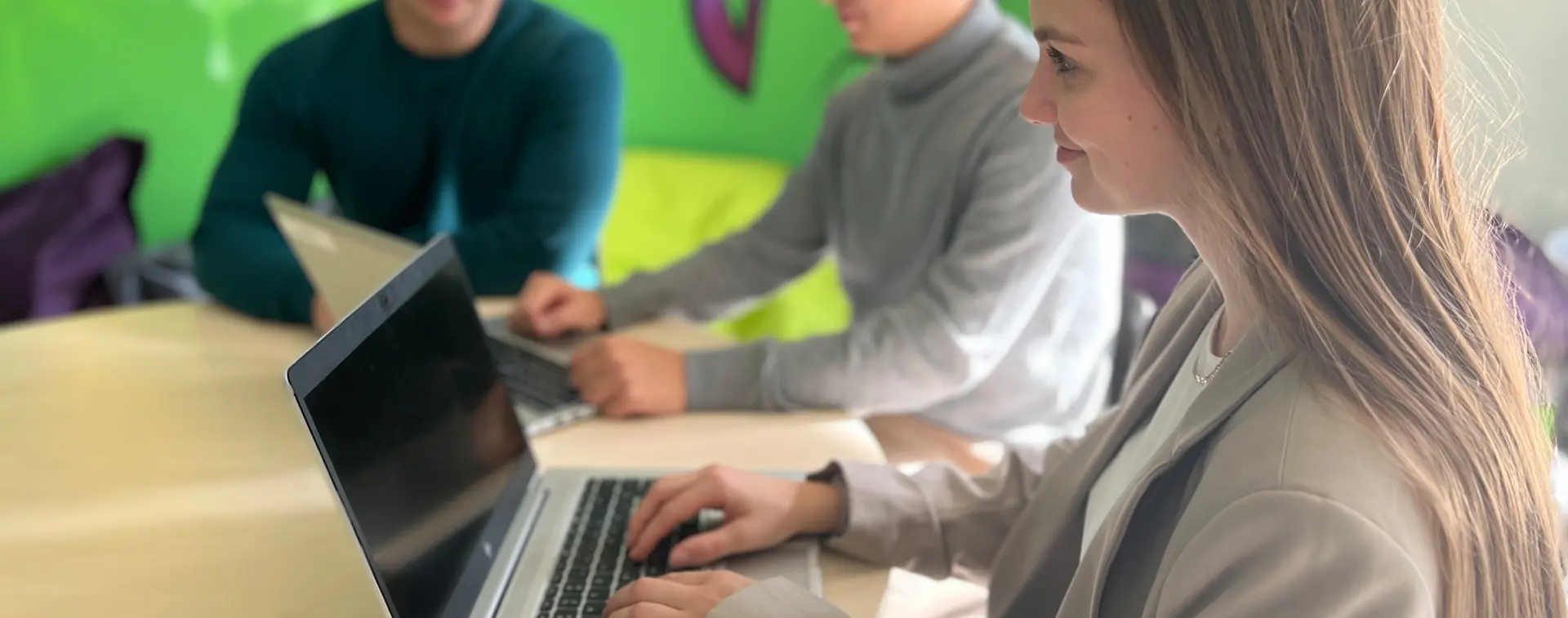 Junge Frau arbeitet an einem Laptop an einem Tisch, im Hintergrund sitzen zwei weitere Personen an Laptops.