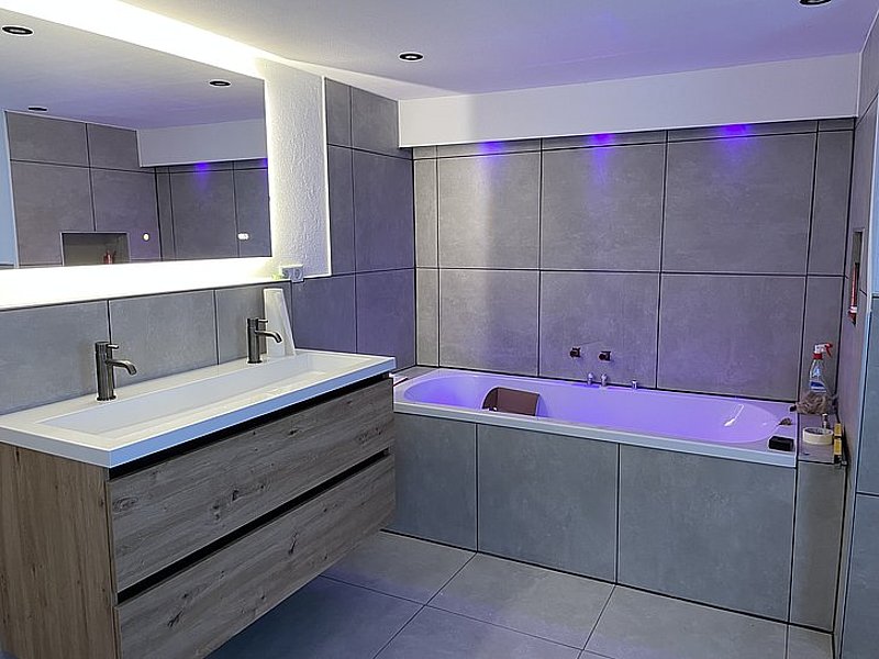 Moderne Badezimmeransicht mit Waschtisch, Badewanne und grauen Fliesen, beleuchtet mit indirektem Licht.