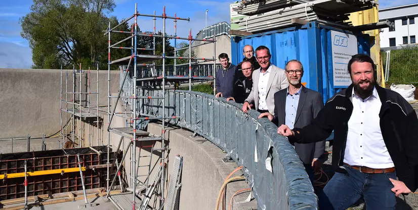 Fünf Personen stehen an einem Baugerüst, umgeben von Baustellenmaterial und einem Container, bei sonnenklarem Wetter.