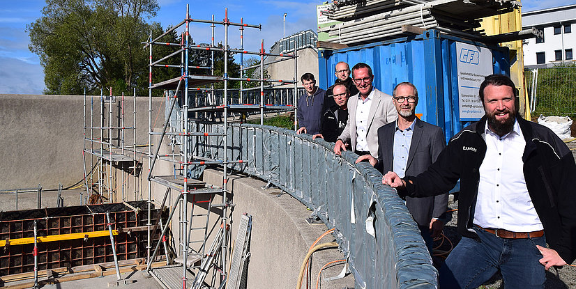 Fünf Personen stehen an einem Baugerüst, umgeben von Baustellenmaterial und einem Container, bei sonnenklarem Wetter.