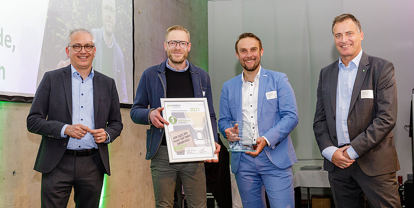 Vier Männer stehen in einem Raum mit Präsentation im Hintergrund, halten Auszeichnungen in den Händen.