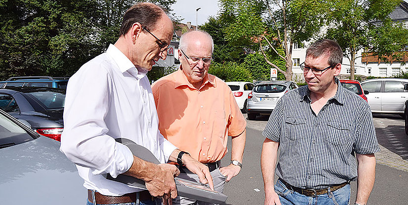 Drei Männer stehen auf einem Parkplatz und diskutieren, während sie ein Dokument in der Hand halten. Autos sind im Hintergrund sichtbar.