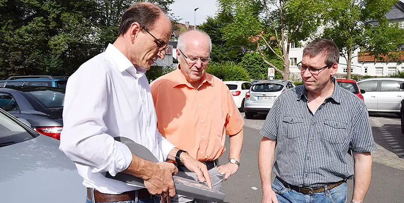 Drei Männer stehen auf einem Parkplatz und diskutieren, während sie ein Dokument in der Hand halten. Autos sind im Hintergrund sichtbar.