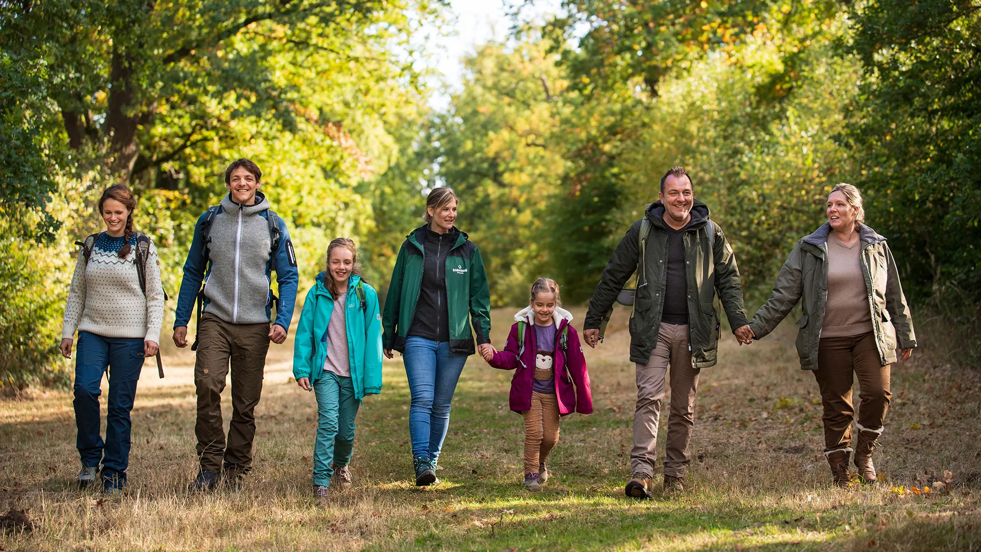 Gruppe von Personen, die gemeinsam auf einem naturbelassenen Waldweg spaziert und dabei Herbstlandschaft, Bäume und Sonnenlicht genießt; Outdoor-Aktivität und Familienwanderung in der Natur.
