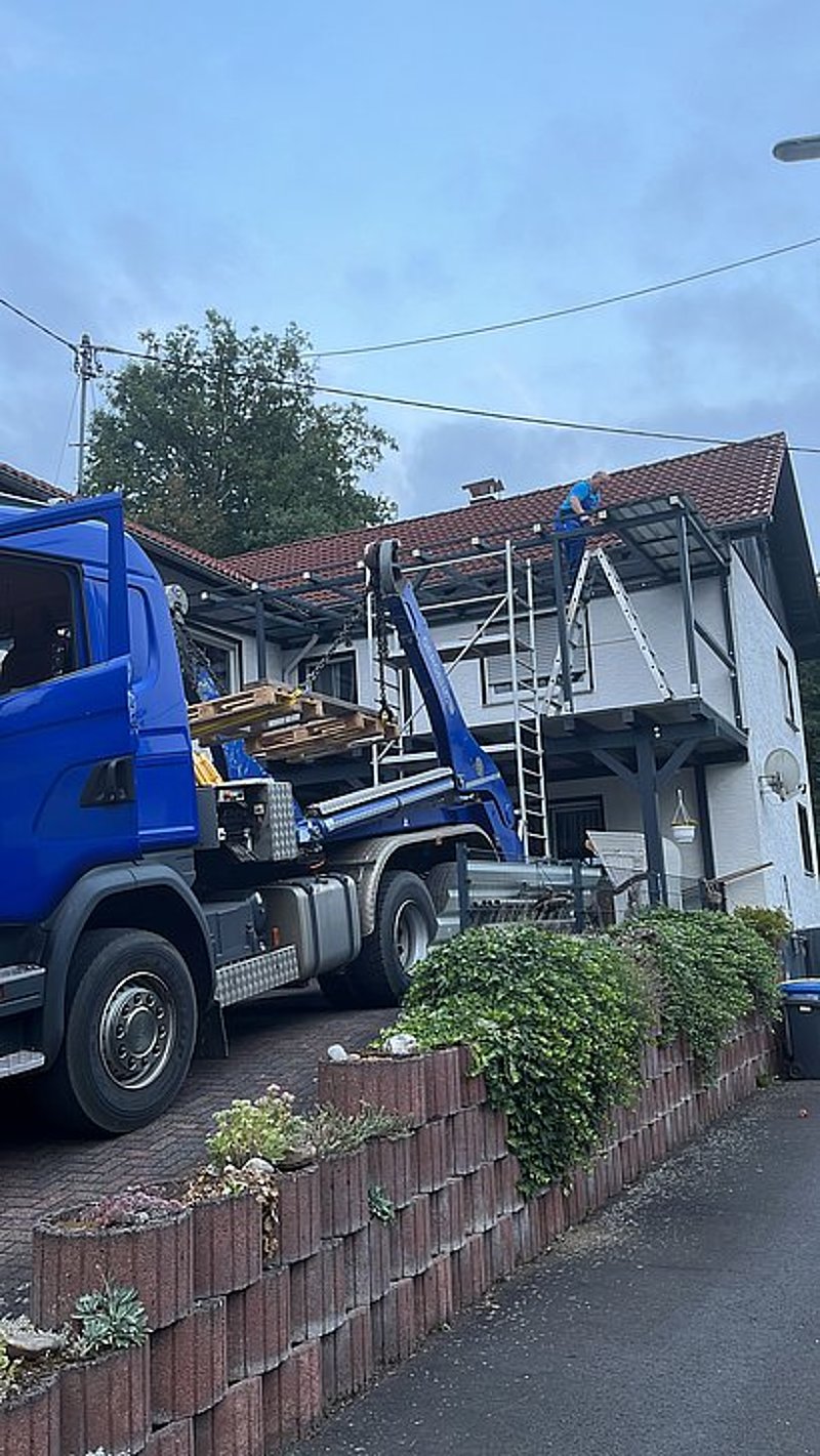 Lkw mit Baumaterial vor einem Haus, während eine Person auf einer Leiter Reparaturen durchführt.