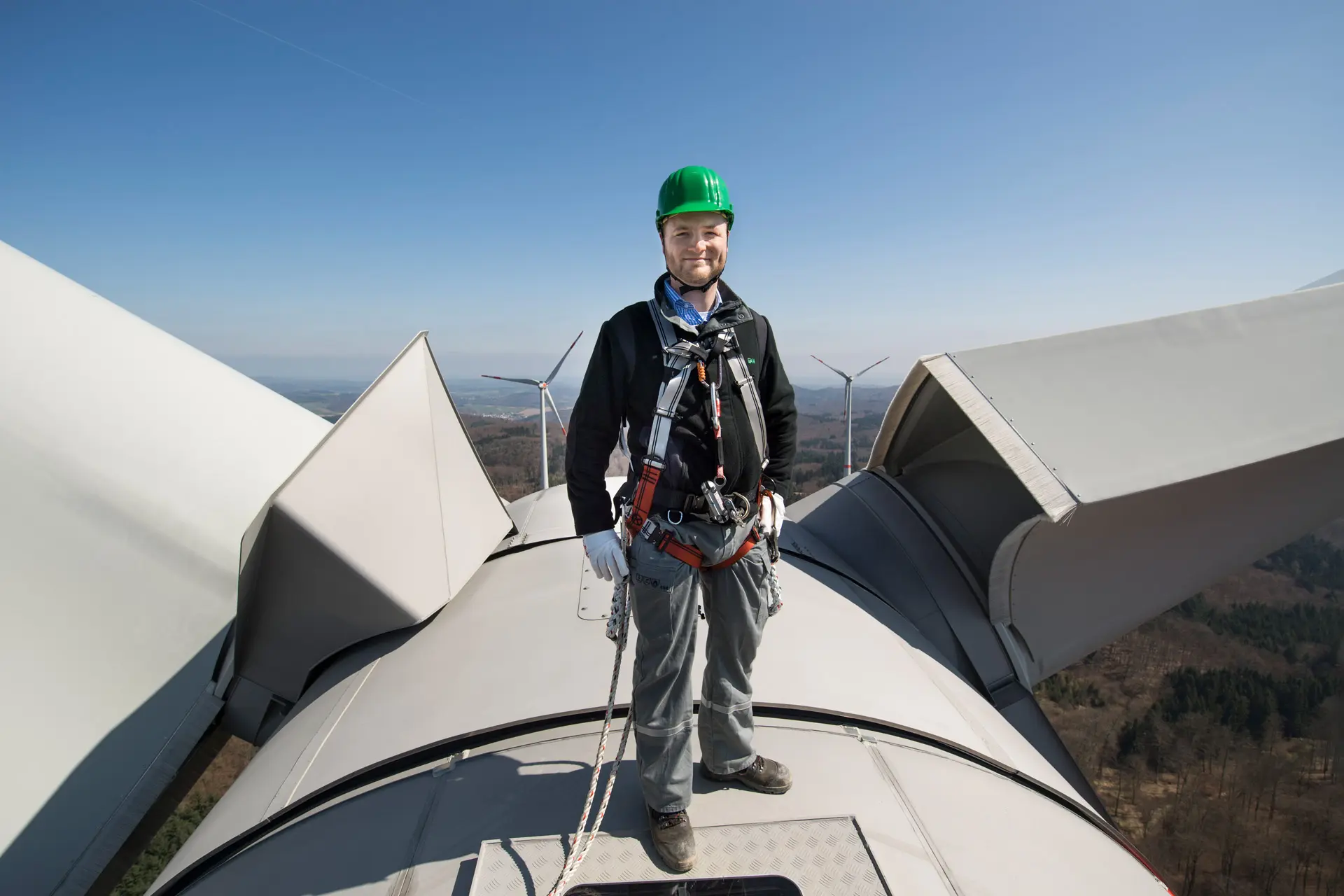 Ein Techniker steht auf einer Windturbine, trägt einen Helm und Sicherheitsausrüstung, mit Blick auf die Landschaft.