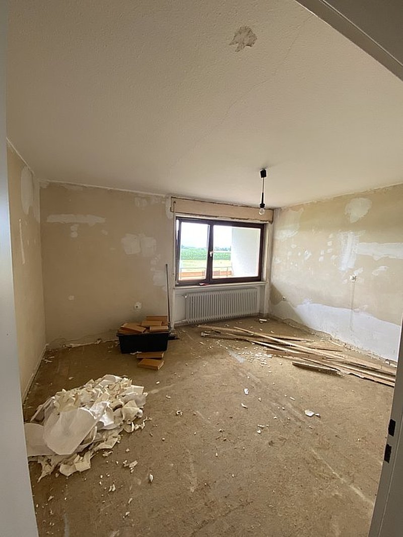 Ein unrenovierter Raum mit einem Fenster, Holzresten und einem Berg aus Sanierungsabfällen auf dem Boden.