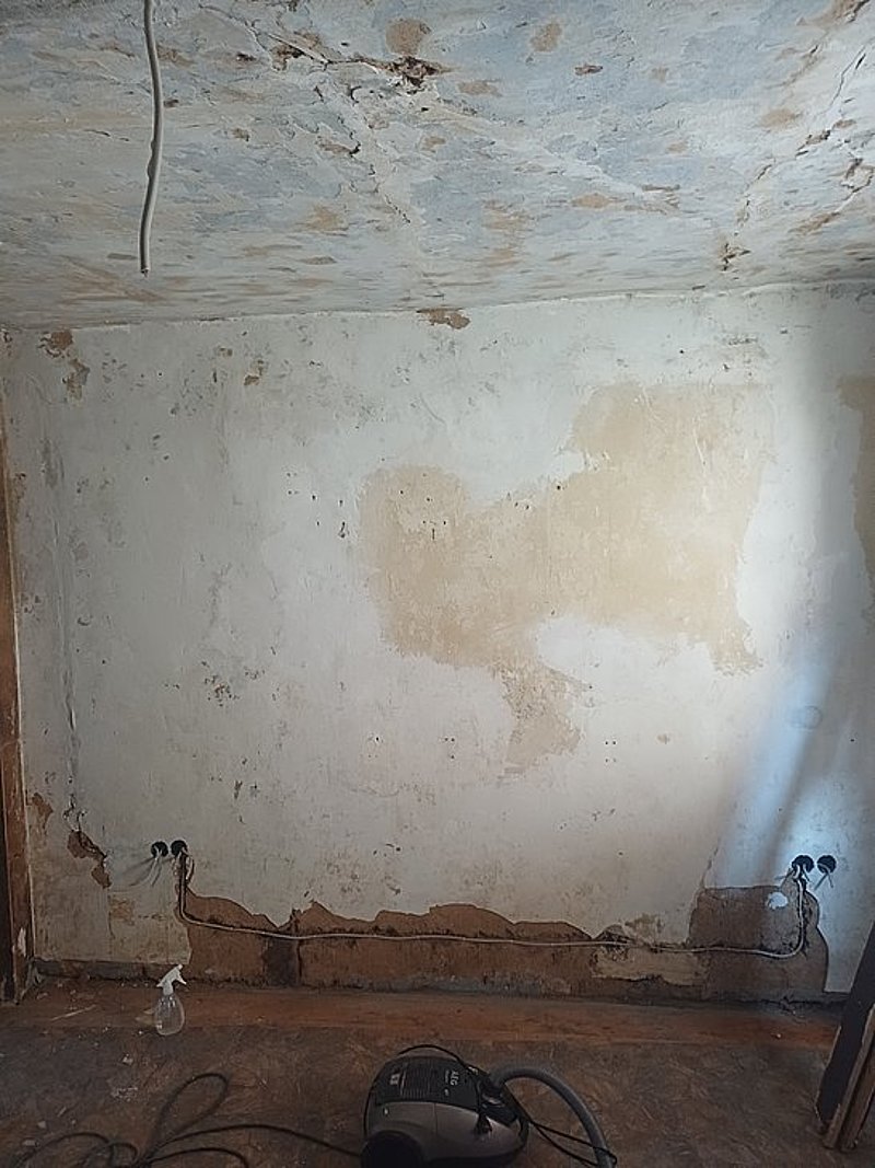 Renovierungsbedürftiger Raum mit unvollständigen Wänden, sichtbarem Boden und einem Staubsauger in der Ecke.