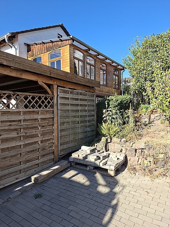 Holzhaus mit großen Fenstern, angrenzender Carport und Gartenbereich mit Pflanzen und Steinen unter blauem Himmel.