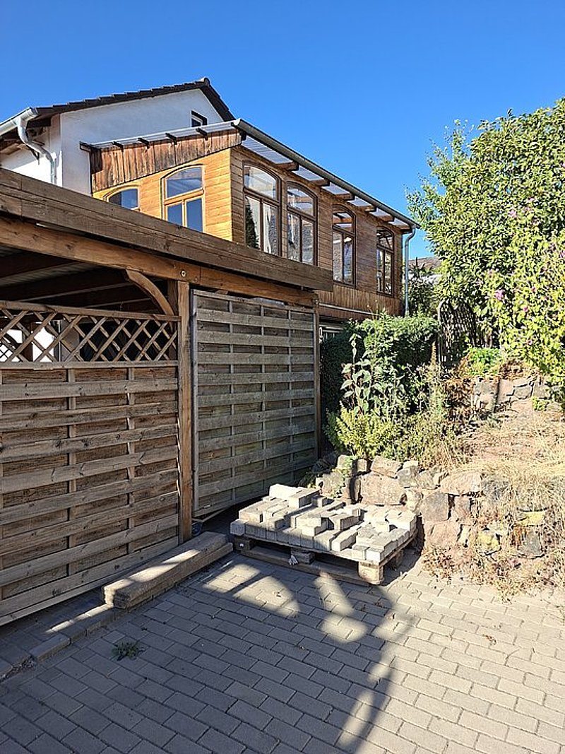 Holzhaus mit großen Fenstern, angrenzender Carport und Gartenbereich mit Pflanzen und Steinen unter blauem Himmel.