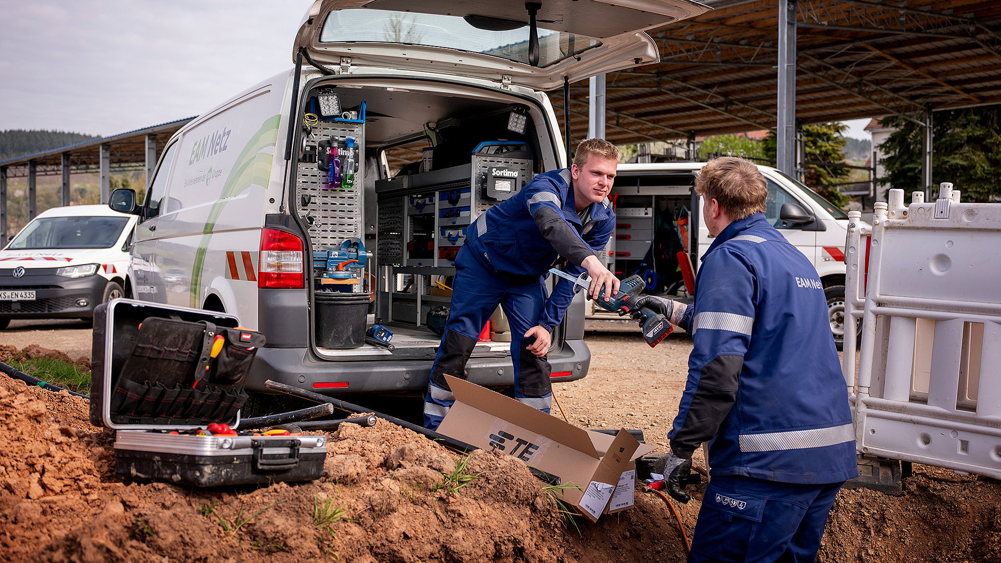 Zwei EAM-Mitarbeiter arbeiten an einem offenen Graben auf einer Baustelle; einer reicht dem anderen ein Werkzeug vor einem geöffneten Servicefahrzeug.