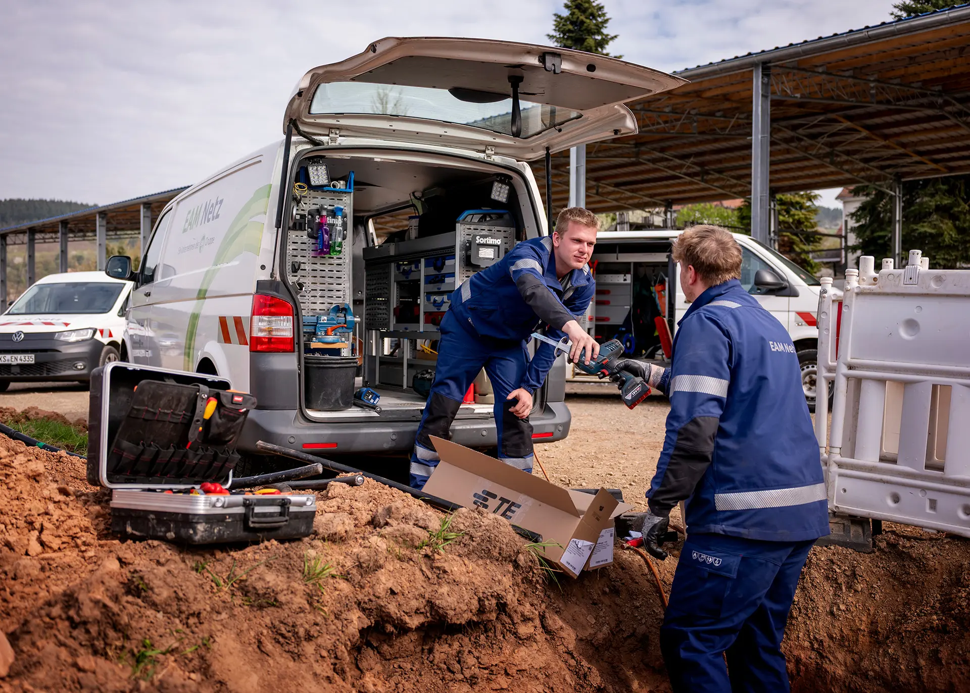 Zwei EAM-Mitarbeiter arbeiten an einem offenen Graben auf einer Baustelle; einer reicht dem anderen ein Werkzeug vor einem geöffneten Servicefahrzeug.
