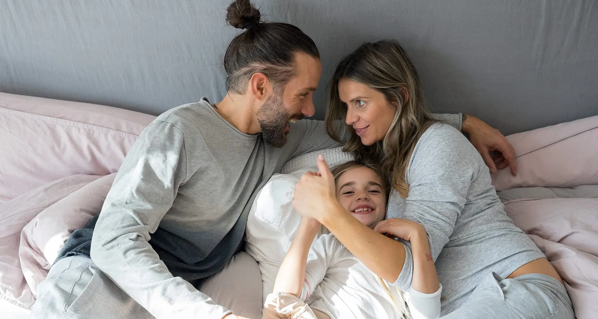  Eine lachende Familie liegt eng zusammen im Bett: Vater mit Dutt und Bart, Tochter und Mutter, die den Daumen hoch hält.