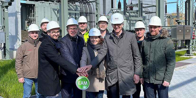 Gruppennfoto von zehn Personen in Schutzhelmen, die gemeinsam einen Knopf drücken, vor einer energie-technischen Anlage.