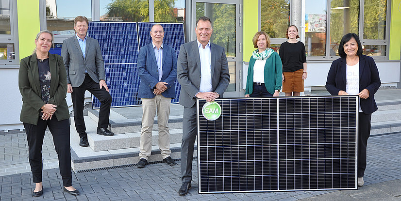 Sieben Personen stehen draußen. Ein Mann in der Mitte hält zwei schwarze Photovoltaikmodule, die mit einem EAM-Logo versehen sind.