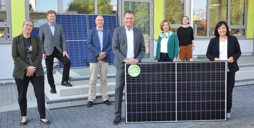 Sieben Personen stehen draußen. Ein Mann in der Mitte hält zwei schwarze Photovoltaikmodule, die mit einem EAM-Logo versehen sind.