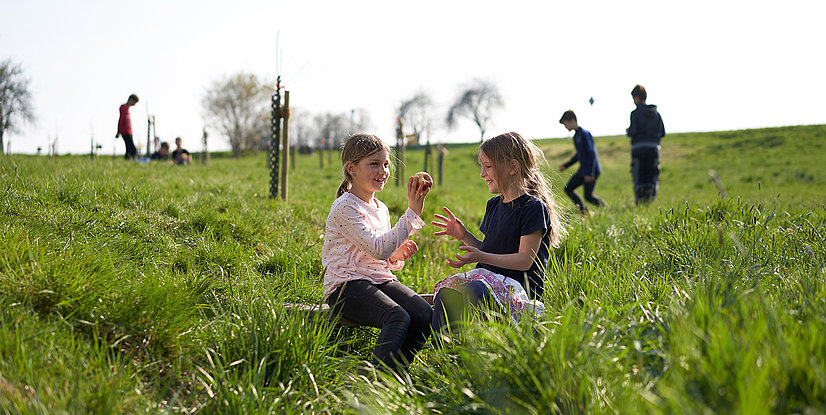 Zwei Mädchen sitzen im Gras und teilen sich einen Apfel. Im Hintergrund sind weitere Kinder sichtbar und spielen..