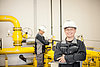 Gasdruckregelanlage Lächelnder Arbeiter in Überalls und Helm vor gelben Rohren in einer Industrieumgebung. Im Hintergrund ein Kollege bei der Arbeit.