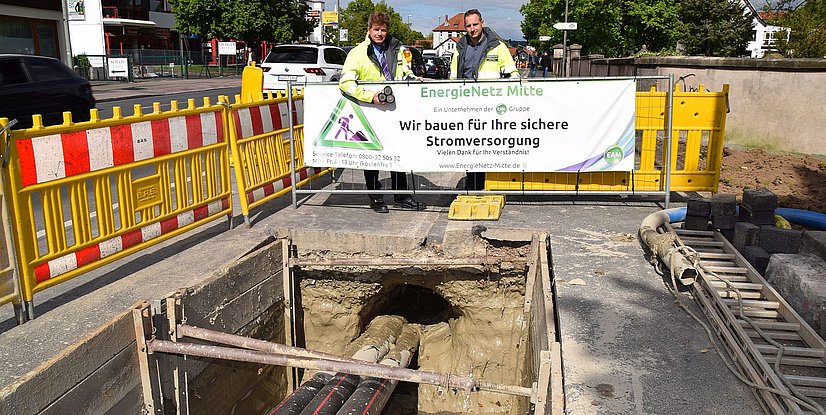 Zwei Arbeiter stehen neben einer Baustelle mit einer Warnsäule und einem Banner zur sicheren Stromversorgung.