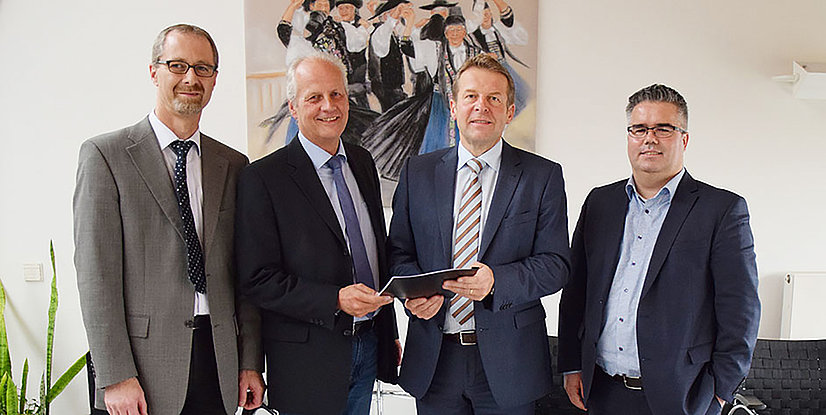 Vier Personen in Anzügen stehen zusammen und halten ein Dokument, vor einem Wandbild mit Tänzern im Hintergrund.