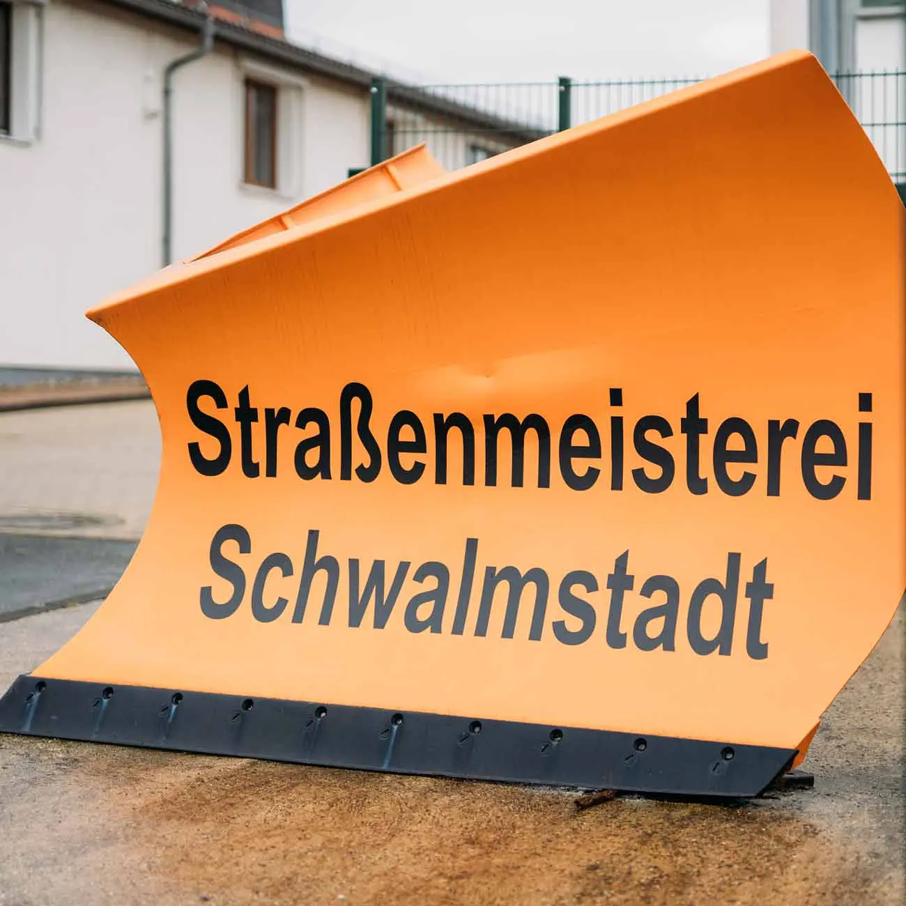 Orangefarbener Schild mit dem Text "Straßenmeisterei Schwalmstadt"" auf einem gepflasterten Untergrund."