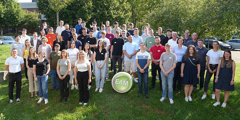 Gruppenfoto von Auszubildenden und dualen Studenten, die freundlich lächeln, vor einer grünen Kulisse mit dem Logo EAM im Vordergrund.