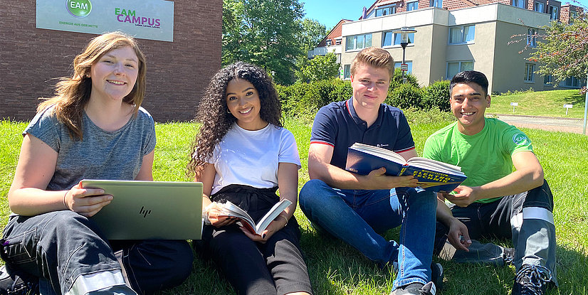 Vier junge Erwachsene sitzen auf einer Wiese, lächelnd und mit Laptops sowie Büchern in der Hand.