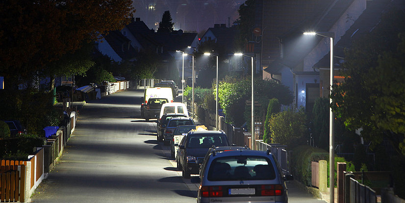 Straßenszene bei Nacht mit beleuchteten Laternen und geparkten Autos in einer ruhigen Wohngegend.